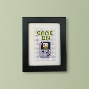 May include: A framed cross-stitch design featuring a pixelated image of a handheld video game console with the text "GAME ON" above it.