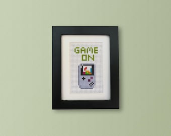 May include: A framed cross-stitch design featuring a pixelated image of a handheld video game console with the text "GAME ON" above it.