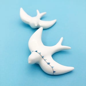 Könnte beinhalten: Zwei weiße Keramik-Vogelfiguren, eine mit einem blauen und weißen Muster, auf einem hellblauen Hintergrund.