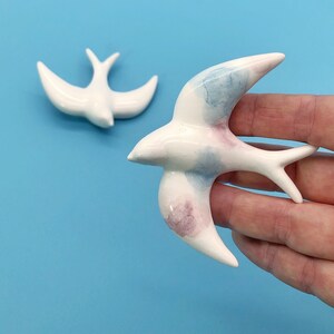 May include: Two ceramic swallow figurines. One is in the background, and the other is held in a hand. The swallows are white with blue and pink watercolor accents on the wings. The background is a solid light blue.