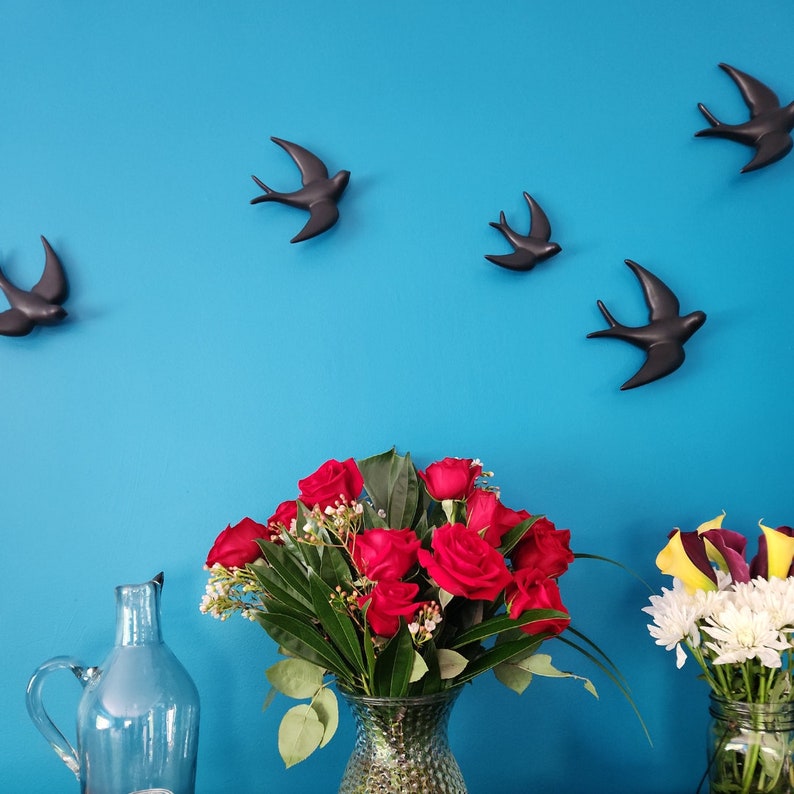 May include: A bouquet of red roses in a clear glass vase with a textured surface. The vase is sitting on a table in front of a blue wall with black silhouette birds.