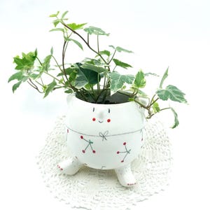 May include: A whimsical white ceramic planter shaped like a bear, with a smiling face and small feet. The planter is decorated with red cherry and blue stitch details. It holds a vibrant green plant with variegated leaves, set on a white lace doily.