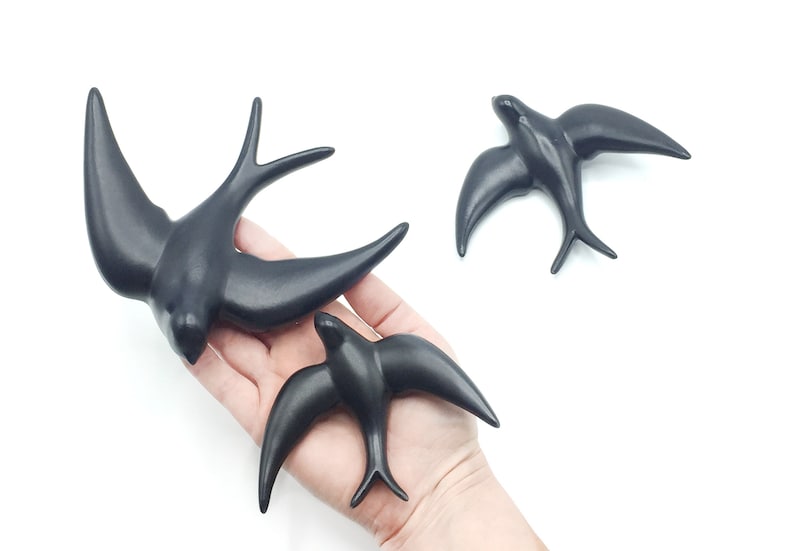 May include: Three black ceramic swallow figurines. The largest swallow is in flight with its wings spread wide. The two smaller swallows are perched with their wings folded.
