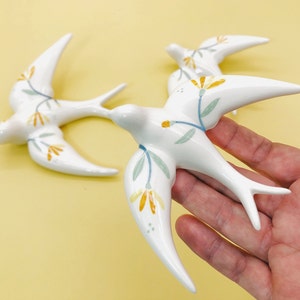 Könnte beinhalten: Zwei weiße Keramikvögel mit floralen Mustern in Gelb-, Blau- und Grüntönen. Die Vögel befinden sich in Flugposition und haben ein zartes, verspieltes Aussehen.