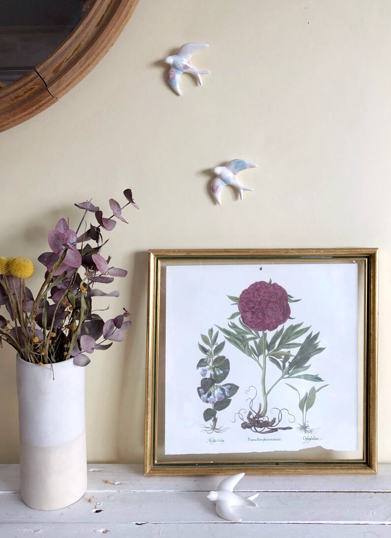 May include: A framed botanical print with a gold frame, featuring a detailed illustration of a peony. Two ceramic swallows with pastel-colored wings are mounted on the wall, and one is on the shelf. A white and beige vase holds dried flowers.