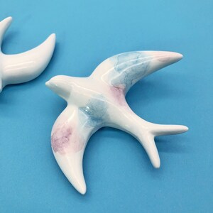 May include: Two ceramic swallow figurines. The birds are white with watercolor-style accents of blue and pink on their wings. The swallows are displayed against a bright blue background.
