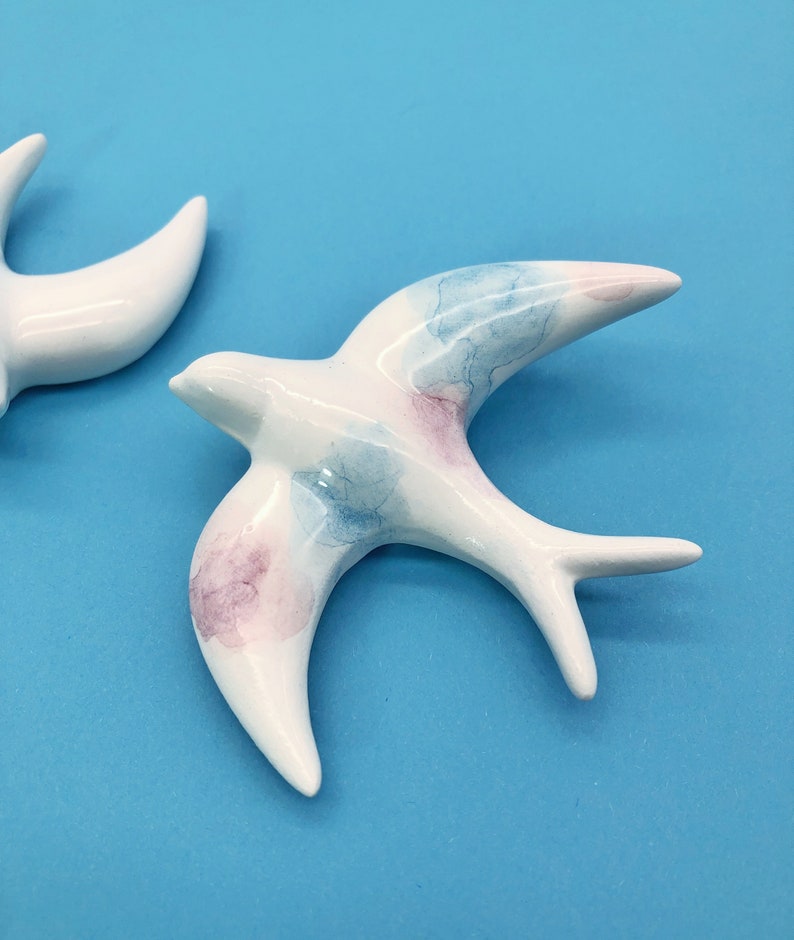 May include: Two ceramic swallow figurines. The birds are white with watercolor-style accents of blue and pink on their wings. The swallows are displayed against a bright blue background.