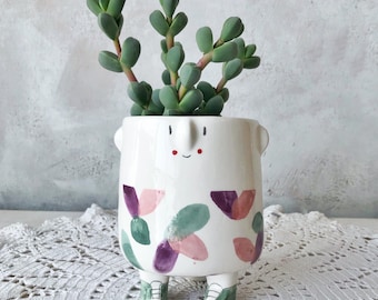 May include: White ceramic planter with a smiling face, pink and green floral pattern, and green shoes. The planter is filled with a succulent plant.