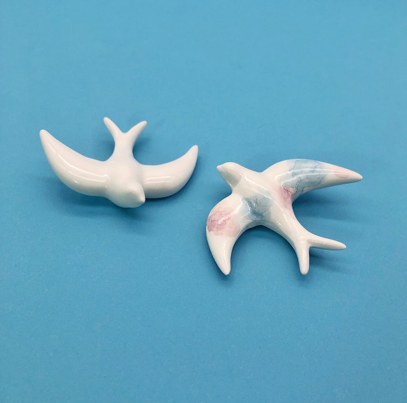 May include: Two ceramic bird figurines. One is solid white, the other features a white base with pink and blue watercolor-style accents. Both have outstretched wings and are set against a solid blue background.