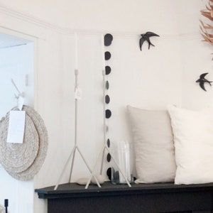 May include: A white wall with a black and white garland of circles, two white metal tripod stands, and two black bird figurines.  There are two pillows on a black shelf.