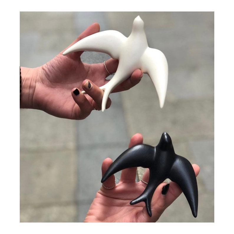 May include: Two ceramic bird figurines, one white and one black, in a flying pose. The figurines are held in the hands of two different people.