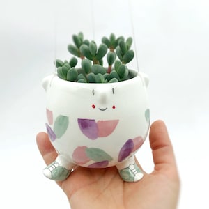 Peut inclure: Un pot de fleurs en céramique blanche avec un visage de dessin animé, contenant une plante succulente verte. Le pot a un motif peint de formes violettes et vertes et de petites chaussures vertes. Le pot est suspendu par de fins fils.