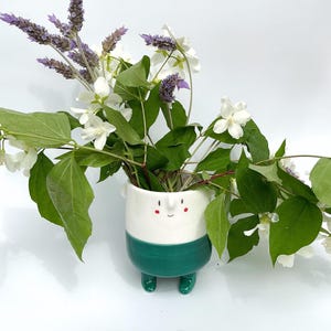 Peut inclure: Un vase en céramique fantaisiste avec un visage souriant, une partie supérieure blanche et une partie inférieure verte avec des pieds assortis. Le vase contient des fleurs blanches, des feuilles vertes et des brins de lavande violets.