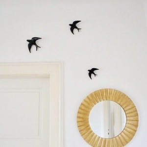 May include: Three black metal swallow bird wall decor pieces are arranged on a white wall. A round, gold-colored mirror with a sunburst design is on the wall below the birds.
