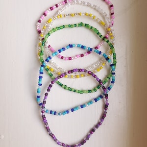 May include: A set of seven colourful beaded bracelets, stacked on top of each other. The bracelets are made of small, round beads in shades of purple, blue, green, yellow, white, and pink.