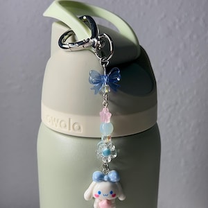 May include: A light green water bottle with a decorative charm. The charm features a blue bow, pink and blue beads, and a white bunny figure with a blue bow. The bottle has the word "owala" printed on it.