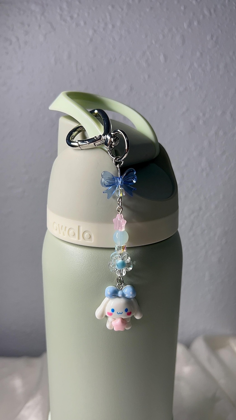 May include: A light green water bottle with a decorative charm. The charm features a blue bow, pink and blue beads, and a white bunny figure with a blue bow. The bottle has the word "owala" printed on it.