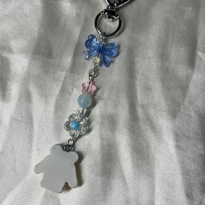 May include: A silver-toned keychain with a heart-shaped clasp. The charm features a blue bow, pink star, blue flower, and a white bear-shaped pendant. The keychain is adorned with clear and blue beads.