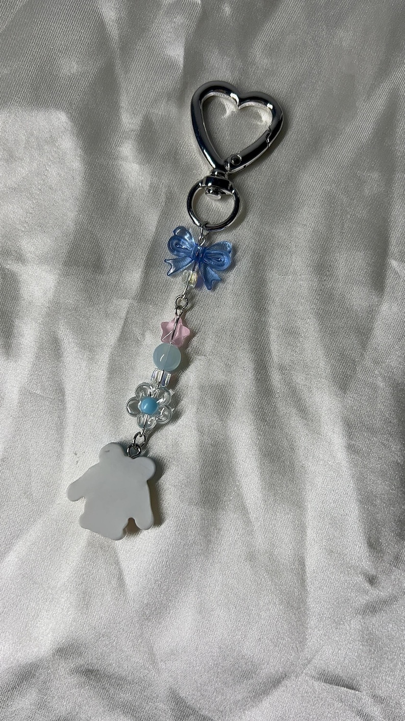 May include: A silver-toned keychain with a heart-shaped clasp. The charm features a blue bow, pink star, blue flower, and a white bear-shaped pendant. The keychain is adorned with clear and blue beads.