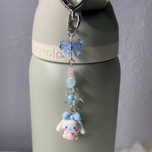 May include: A silver keychain with a light blue butterfly charm, pink and blue beads, a flower charm, and a white cartoon character with a blue bow. The keychain is attached to a green water bottle.