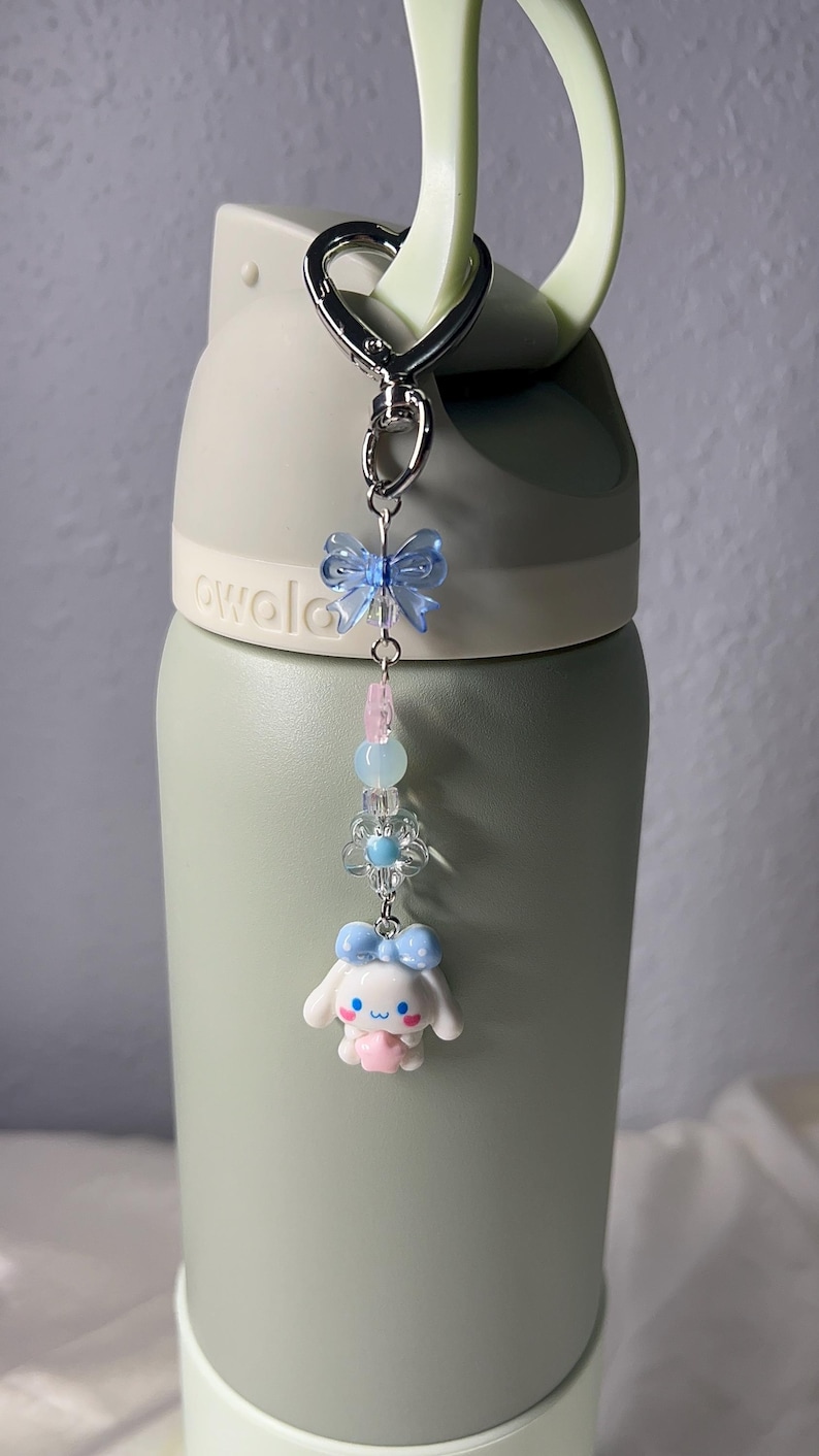 May include: A silver keychain with a light blue butterfly charm, pink and blue beads, a flower charm, and a white cartoon character with a blue bow. The keychain is attached to a green water bottle.