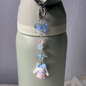 May include: A decorative keychain with a silver clasp, featuring a blue bow, pink star, and a white cartoon character with a blue bow. The keychain is attached to a green water bottle.