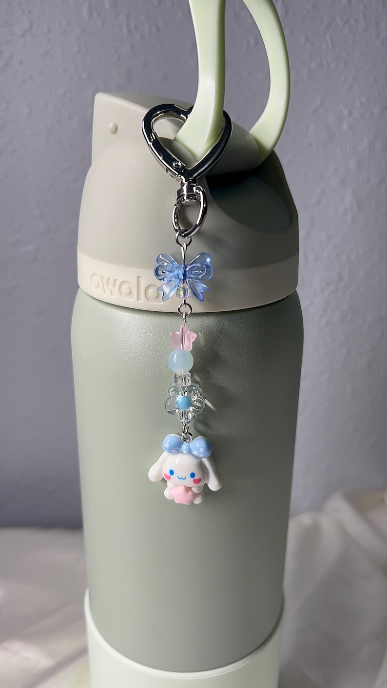May include: A decorative keychain with a silver clasp, featuring a blue bow, pink star, and a white cartoon character with a blue bow. The keychain is attached to a green water bottle.
