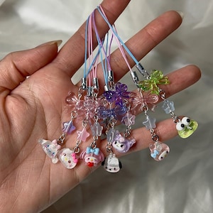 May include: A collection of colorful phone charms featuring various designs, including flowers, stars, and cartoon characters. Each charm is attached to a pastel-colored cord with a silver-tone clasp. The charms are made of clear and translucent materials.