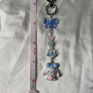 May include: A silver keychain with a heart-shaped charm and a decorative charm featuring a white cartoon character with a blue bow, pink star, and other pastel-colored beads. A pink and white measuring tape is on the left.