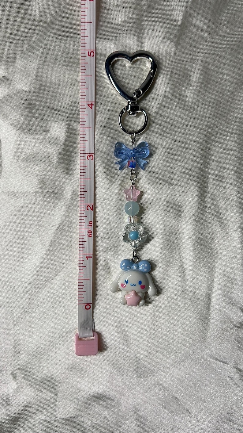 May include: A silver keychain with a heart-shaped charm and a decorative charm featuring a white cartoon character with a blue bow, pink star, and other pastel-colored beads. A pink and white measuring tape is on the left.