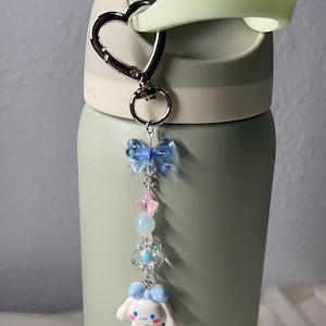 May include: A light green water bottle with a heart-shaped carabiner clip holding a charm. The charm features a blue bow, pink and blue beads, a flower, and a white cartoon character with a blue bow.