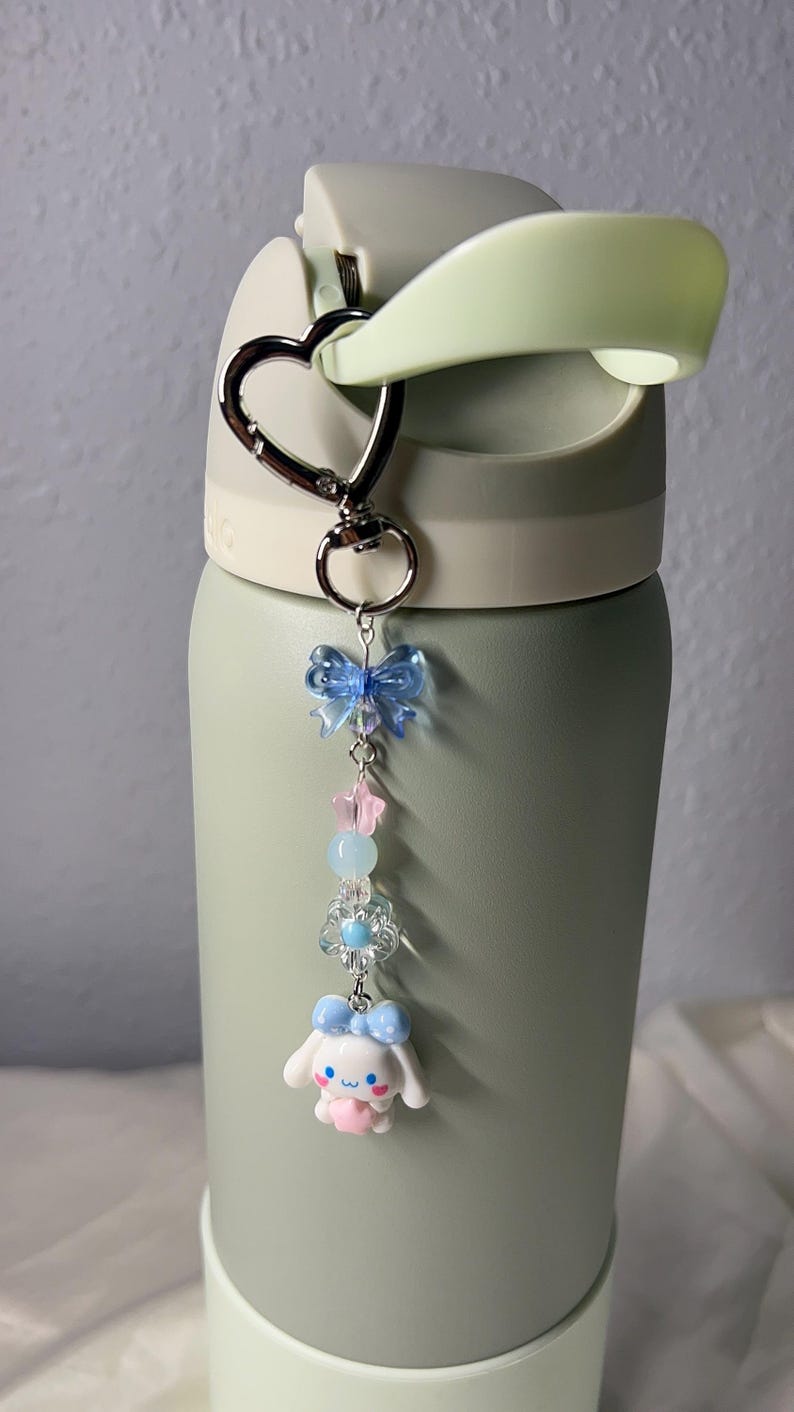 May include: A light green water bottle with a heart-shaped carabiner clip holding a charm. The charm features a blue bow, pink and blue beads, a flower, and a white cartoon character with a blue bow.