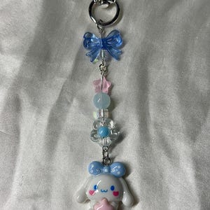 May include: A silver heart-shaped keychain with a charm featuring a white cartoon character holding a pink star. The charm is adorned with blue bows, pink stars, and clear and blue beads.