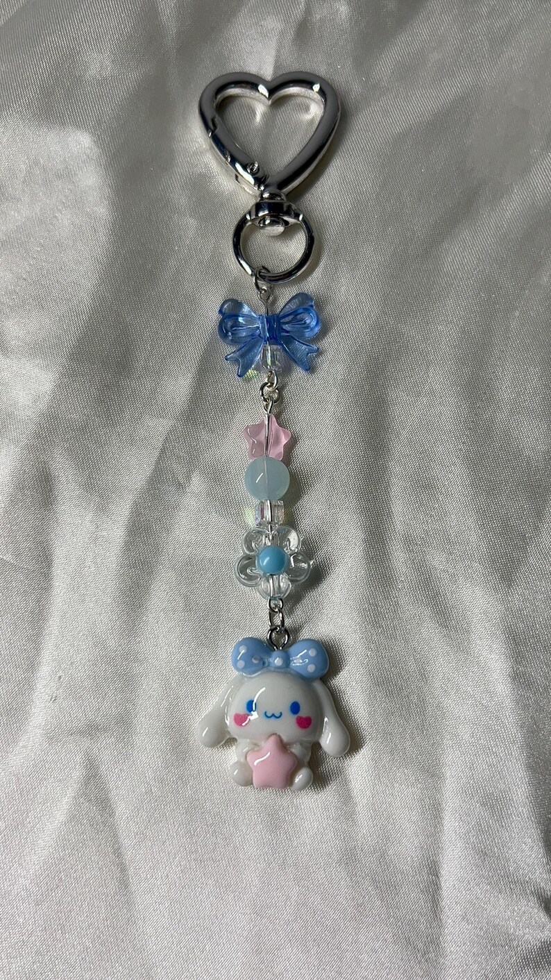 May include: A silver heart-shaped keychain with a charm featuring a white cartoon character holding a pink star. The charm is adorned with blue bows, pink stars, and clear and blue beads.