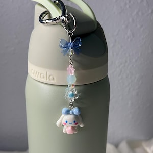 May include: A light green water bottle with a decorative charm. The charm features a blue bow, pink stars, clear beads, a flower, and a white cartoon character with a blue bow. The bottle has a light green handle and a silver carabiner clip.