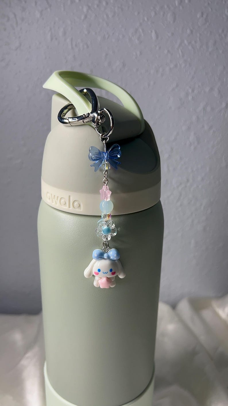 May include: A light green water bottle with a decorative charm. The charm features a blue bow, pink stars, clear beads, a flower, and a white cartoon character with a blue bow. The bottle has a light green handle and a silver carabiner clip.