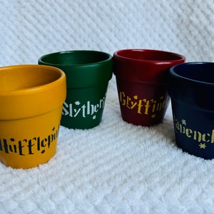 May include: Four small terracotta flower pots painted in the colours of the Hogwarts houses. The pots are labelled "Hufflepuff", "Slytherin", "Gryffindor", and "Ravenclaw".