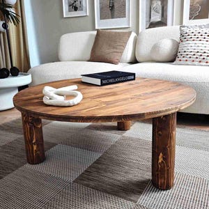 Puede incluir: Una mesa de centro redonda de madera con un acabado marrón oscuro. La mesa tiene cuatro patas cilíndricas. Un nudo decorativo blanco y un libro titulado "MICHELANGELO" descansan sobre la mesa. La mesa está sobre una alfombra estampada.