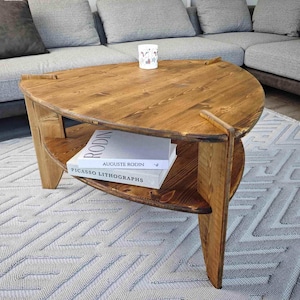 Może przedstawiać: Trójkątny drewniany stolik kawowy z dolną półką. Stół ma ciepłe, brązowe wykończenie i unikalny kształt. Książki są umieszczone na dolnej półce. Kubek stoi na powierzchni stołu. Stół stoi na wzorzystym dywanie.