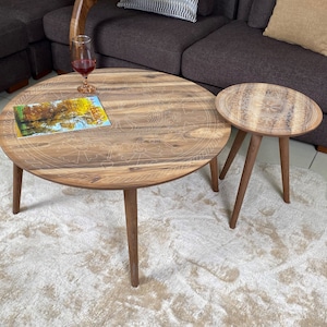 May include: Two round wooden coffee tables with intricate carved designs. The larger table has a magazine on top and a glass of wine. The smaller table is next to the larger table.
