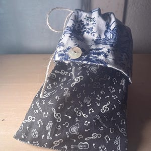 May include: A drawstring bag with a blue and white toile pattern on the top and a black fabric with white musical notes on the bottom. The bag is closed with a button and a natural twine drawstring.