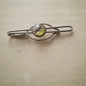 May include: Silver tie clip with a round glass cabochon featuring a painted scene of horses racing.
