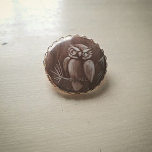 Op de afbeelding: Een gouden broche met een bruine cameo met een witte uil die op een tak zit.