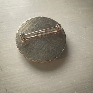 Op de afbeelding: De achterkant van een ronde, goudkleurige broche met een decoratieve rand. De broche heeft een pin en een sluiting.