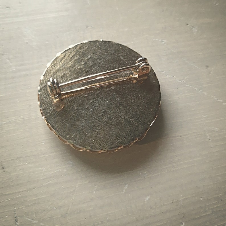 Op de afbeelding: De achterkant van een ronde, goudkleurige broche met een decoratieve rand. De broche heeft een pin en een sluiting.