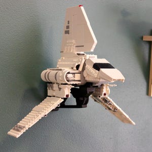 Puede incluir: Un modelo de construcción de ladrillos de plástico blanco de una nave espacial con un estabilizador vertical alto, dos alas en ángulo y una ventana de cabina negra. El modelo está montado en un soporte de pared y muestra un diseño exterior detallado.