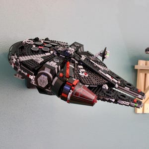 Könnte beinhalten: Ein schwarzes und graues Modellraumschiff, aus Bausteinen konstruiert, ist an einer hellblauen Wand befestigt. Das Modell hat rote und silberne Akzente mit detailliertem Design. Ein weiteres ähnliches Modell ist auf einem Holzregal zu sehen.