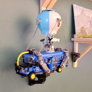 Peut inclure: Un modèle Lego bleu et blanc d'un bus suspendu par une montgolfière. Le bus a des roues jaunes et un tuyau jaune. Le ballon est bleu et blanc. Le modèle est monté sur un mur.