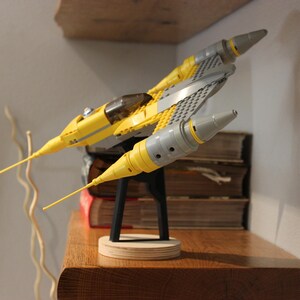 May include: A yellow and gray Lego Star Wars V-4X-D starfighter model on a black stand. The model is on a wooden surface.
