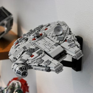 Puede incluir: Un modelo de nave espacial Lego Millennium Falcon gris y blanco montado en un soporte de pared negro. El modelo tiene detalles rojos y un motor gris y negro.